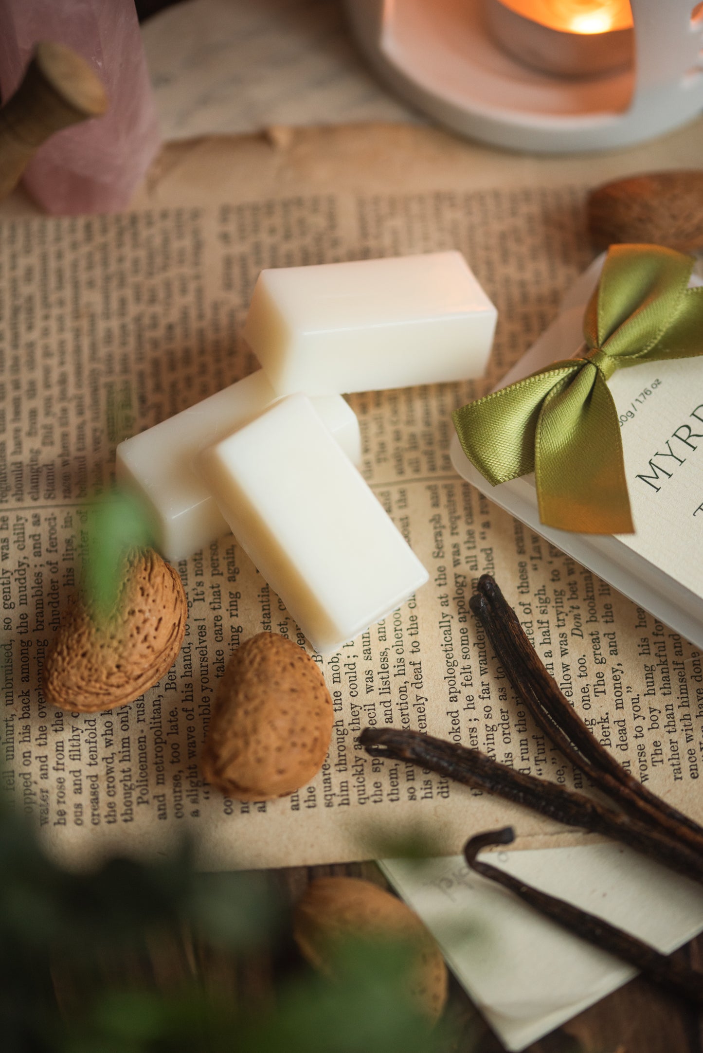 White rectangular wax bars with a green bow, almonds, vanilla beans, and a candle on a textured surface.